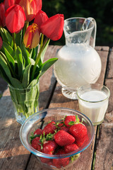 Fresh strawberries on plate on old wooden background. Red tulips in vase on table. Jar and glass of milk. Breakfast time. Healthy food. Vegetarian dish. Spring time
