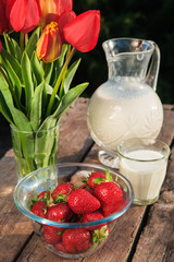 Fresh strawberries on plate on old wooden background. Red tulips in vase on table. Jar and glass of milk. Breakfast time. Healthy food. Vegetarian dish. Spring time
