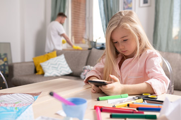 Cute little girl scrolling through photos in smartphone while sitting by table