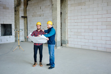 Two young builders in protective helmets discussing sketch in blueprint