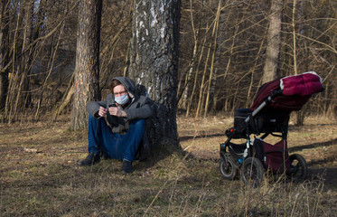 A young man violated quarantine and walks with a stroller in the park. A young father is sitting under a tree with a phone, instead of walking with a child.