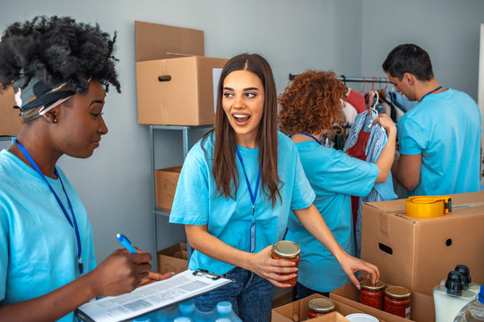 Small Mixed Race Group Of People Working In Charitable Foundation. Male And Female Teenage Food Bank Volunteers Sort Canned Food Items In Cardboard Boxes. Happy Volunteer In Community Food Bank