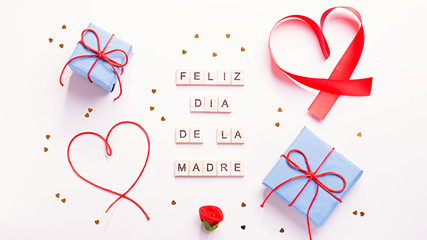 Happy Mother's Day greeting card with red hearts, blue gift boxes and golden confetti on pastel table top, overhead shot. Feliz dia de la madre text that mean Happy Mothers day - obrazy, fototapety, plakaty