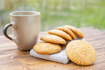 Hot sesame round cookies, homemade healthy snack. Honey bars with sesame seeds on wooden board, cup with tea or coffee. Plant background, beautiful breakfast outdoor. Natural light, copy space.