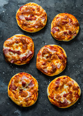 Mini ham, tomato sauce, mozzarella, sun dried tomatoes pizza on a dark background, top view. Delicious breakfast, snack, tapas