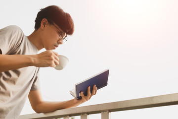 Fototapeta premium Young handsome asian man reading book and drinking tea at room balcony in free time from working at home, Knowledge and learning concept.