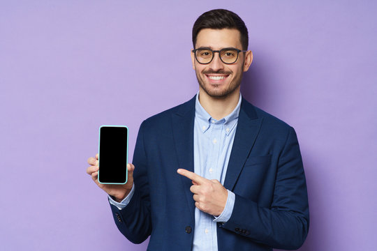 Young businessman smiling to viewer, presenting blank screen cellphone, promoting new app on copy space, looking confident, isolated on purple background