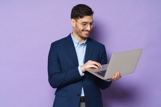 Young businessman chatiing with mates through video call, holding open laptop in hands, smiling happily, looking at screen through eyeglasses, isolated on purple background