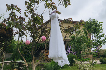 Wedding white long dress hanging on a hanger, on a tree in a flowering garden. The morning of the bride. Photography, concept.