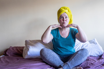 A young woman sits on a bed and does gymnastics for her face. The concept of quarantine and...