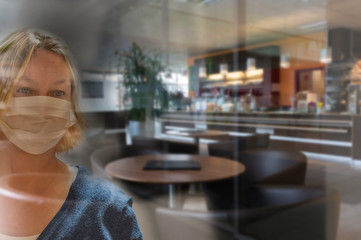 frau mit mundschutz schaut durch eine glasscheibe  in ein geschlossenes cafe