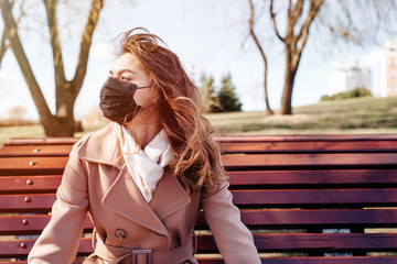 Young woman wearing a face mask as a protection from air disease, allergens or pollution sitting on bench in the park