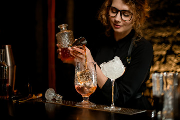 Beauty young female bartender gently pours alcoholic drink from jigger into large glass with ice