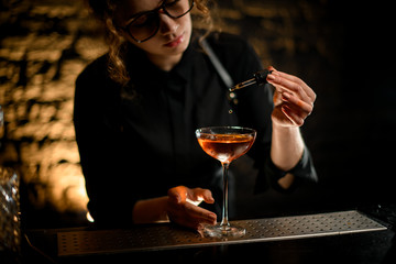 Young female barman adding ingredient to glass with drink