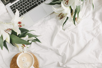 Comfortable bedroom with laptop, coffee cup and flowers on the white bed sheet. Good morning and weekend concept in flat lay style