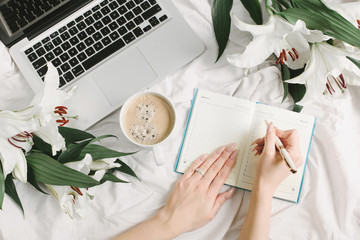 Woman write in opened notebook, laptop, coffee cup and flowers on the white bed sheet. Good morning...