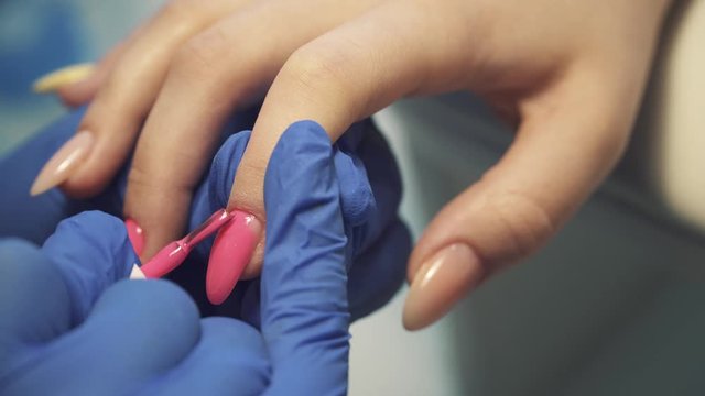 Paints female nails with pink gel polish in blue protective gloves