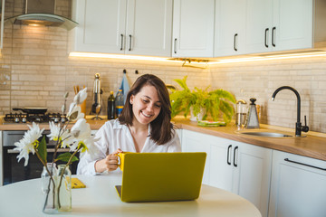 woman drinking tea from yellow mug working on laptop. kitchen on background