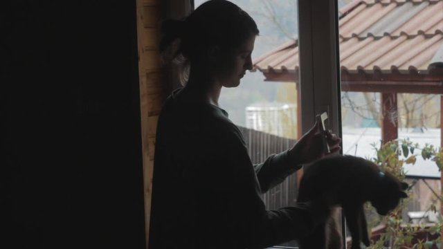 Silhouette of a girl opens a window and takes a breath of fresh air. Female stays at home during quarantine. Woman keeps social distancing. Cat sitting on a windowsill.