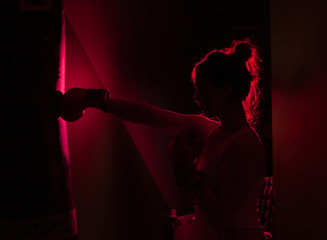 Silhouetted female boxer with boxing gloves punch a punching bag in red neon light over dark background