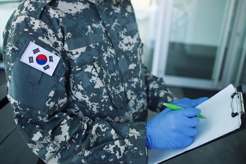 Korean soldier in camouflage uniform with rubber gloves, mask guarding in front of hospital's door controlling who gets in or out. Korea military helping in fight against coronavirus or COVID-19.