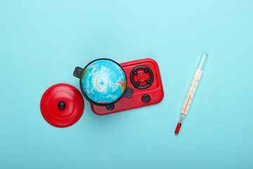 Global warming concept. Mini globe in toy pan on stove and thermometer. Blue pastel background. Top...