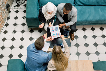 Top view of four young business people of different nationalities working with financial documents and looking together at graphs and charts, sitting around table in modern loft office meeting room