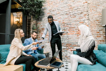 Handsome african businessman gives working papers to his multiethnic team of businesspeople during the meeting for discussing financial reports and company development, in modern loft room