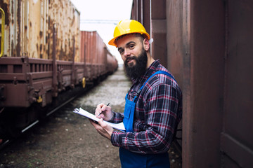 Checking on cargo arrived via freight trains. Railroad worker with clipboard standing by the shipping containers and looking to the camera. Keeping track of arrived cargo.