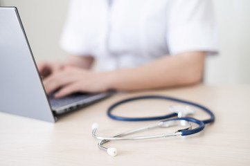 Unrecognizable female doctor in uniform uses a laptop. Faceless medical worker writes a report on the computer at the desk. Cropped.