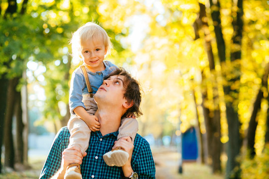 Cute Handsome Dad Carrying Boy On Back In Park, Smiling. Dad Giving His Young Son A Piggy Back Ride As They Both Laugh With Pleasure And Enjoyment, Low Angle Against  Autumn Park With Copyspace