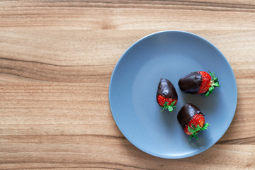 Top view of fresh healthy strawberries dipped in dark chocolate on a blue plate with a wooden background