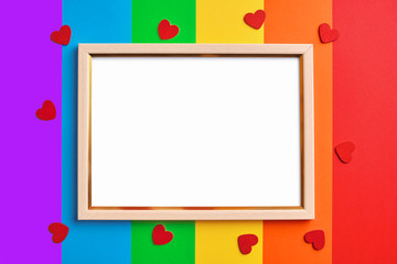 wooden frame with blank white copy space on multicolored rainbow flag background and little red hearts. Pride day concept, flat lay