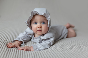 A cute little girl in a light beautiful light bodysuit and a cap on the bed at home looks at the camera. Portrait of a cute child lying on a blanket. Space for text.