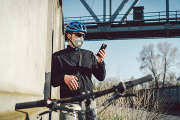 The courier in protective mask on bicycle stopped and was watching the phone application. Delivery service in quarantine. A man in a respirator uses a mobile phone. Bicycle messenger works