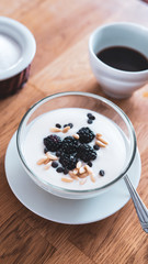 breakfast on a table, set with a cup of coffee and a natural yogurt with blackberries, chocolate chips and pine nuts on top