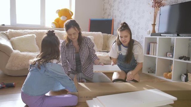 A mother and two daughters together unpack a box with a new rack for the living room.