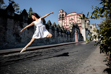 Obraz premium Young girl extend her arms and legs when dancing and jumping wearing flats ballet and white dress in old and empty street of México in front of old church under a blue sky on a spring morning.