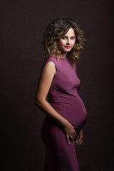 beautiful pregnant woman in dress, hairstyle and beautiful makeup looks forward . Photo taken in a photo studio on a brown background texture. He has a smile and joy in his eyes