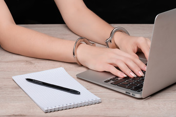 Hands cuffed on the keyboard of a laptop against the background of a Notepad with a pen. The concept of restricting journalistic activity or freedom of speech.