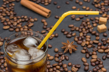 Glass of iced coffee, yellow straw. On black background, coffee grains, cinnamon stick, star anise.