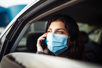 Fototapeta na wymiar Woman wearing a medical sterile mask in a taxi car on a backseat looking out of window talking on the phone. Girl passenger waiting in a traffic jam during coronavirus quarantine. Healthcare concept.