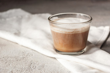 A glass of morning coffee with milk and milk foam, on a white napkin on a concrete table. Cozy warm tint, copy space