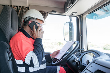 Truck driver with helmet, safety glasses and high visibility clothing talking on the phone and with a folder reviewing notes