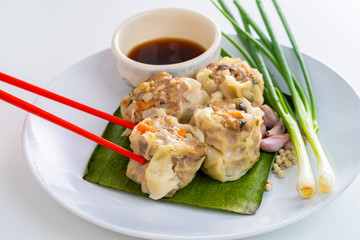 Close up Chopsticks Picking Asian Steam  Dumplings or Dim Sum a Famous Chinese Food with Pork and Shrimp on Banana Leaf and White Dish with Garlic, Spring Onion and Peppers on Isolated Background.