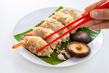 Close up Chopsticks Picking Asian Steam  Dumplings or Dim Sum a Famous Chinese Food with Pork and Shrimp on Banana Leaf and White Dish with Garlic, Spring Onion and Peppers on Isolated Background.