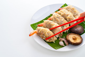Close up Chopsticks Picking Asian Steam  Dumplings or Dim Sum a Famous Chinese Food with Pork and Shrimp on Banana Leaf and White Dish with Garlic, Spring Onion and Peppers on Isolated Background.