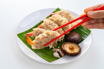 Close up Chopsticks Picking Asian Steam  Dumplings or Dim Sum a Famous Chinese Food with Pork and Shrimp on Banana Leaf and White Dish with Garlic, Spring Onion and Peppers on Isolated Background.