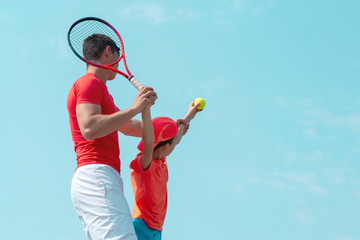 A young tennis coach or instructor teaches a child a serving technique. Boy tennis player throws the ball up for serve. Kids sports school. Blue sky background. Banner. Copy space for text.