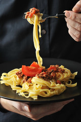 In a man’s hand is a black plate with pasta bolognese.  In the other hand is a fork with pasta.  Close-up.  The vertical orientation of the frame.  Black background.